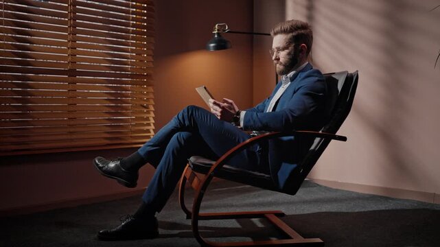 Side view of man in business suit sitting on chair. Professional man taping tablet screen at dim room. Businessman wearing official suit and glasses. Man occupied by office lamp and flowerpot.