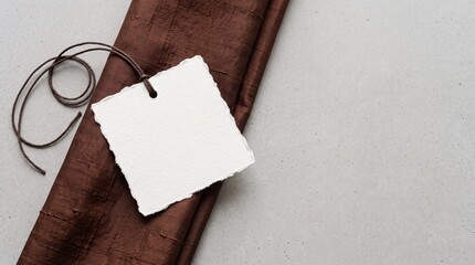 Fototapeta premium A blank white tag with a rough edge is tied with a brown string. The tag rests on a brown fabric surface, creating a simple and elegant composition.