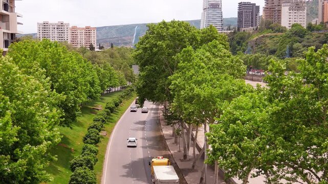 High-angle view of cars driving on a road along the mtkvari river embankment in tbilisi, georgia, with a lush green park on one side and modern city buildings in the background