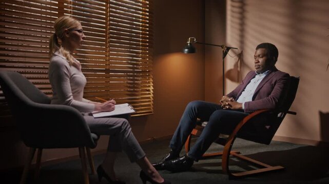 Actor sitting in front of film producer. Male actor auditioning for role in movie. African American communicating with casting director. Female director conducting audition. Dark room, window blinds.
