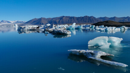 Floating blue icebergs in a calm glacial lagoon under clear skies in Iceland with a mountain backdrop © Martin