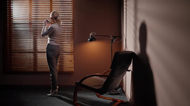 Casting manager having work break. Female standing near window and looking through blinds. Woman wearing heels standing near office chair and lamp. Female drinking coffee in dark room.