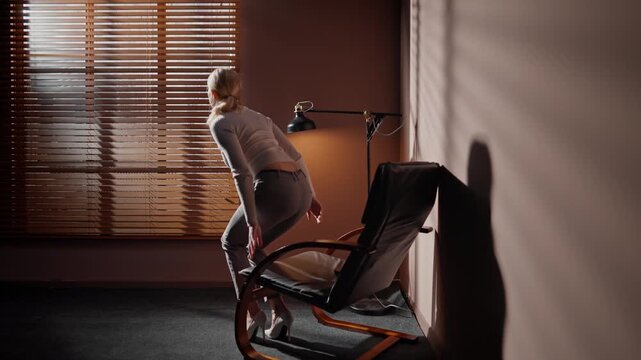 Woman putting away tablet. Female sitting on office chair near lamp. Woman standing up and going to window. Professional woman looking through window blinds. Woman isolated at dark sunny room.