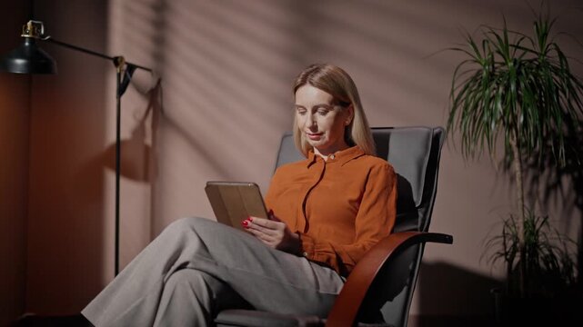 Woman working late in evening. Female sitting on chair in dim room. Woman working with tablet. Female isolated in dark loft room. Woman in bright orange shirt. Female isolated in cosy room.
