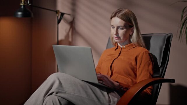 Woman sitting in front of laptop. Female sitting on chair holding laptop. Woman working isolated in modern room. Female using laptop at background of lamp. Woman in orange shirt with crossed legs.