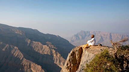 Fototapeta premium Vast rocky mountain valley at sunset with a lone traveler seated on a cliff edge, overlooking rugged layered ridges for desert travel and nature tourism