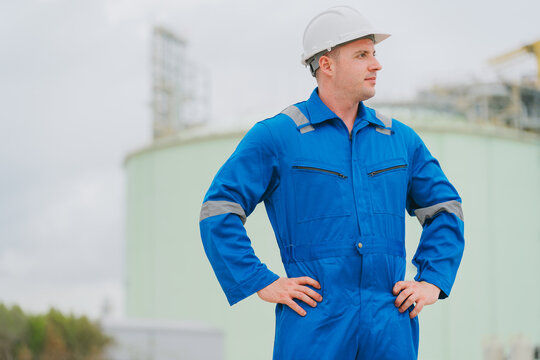 Oil industry concept. Industrial worker in blue coverall and hardhat stands with hands on hip at storage tank