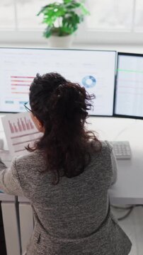 Woman analyst reviews spreadsheet data report on computer. Three monitors show accounting and finance figures in a clean office, with charts and KPIs. Clear data driven financial audit concept.