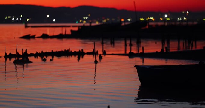 Silhouette of fishing boat and wooden posts in calm sea at vivid orange sunset and night city lights as background