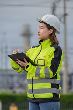 Professional female industrial engineer wearing safety gear uses a clipboard to inspect electrical infrastructure at a modern power plant facility to ensure reliable daily energy production.