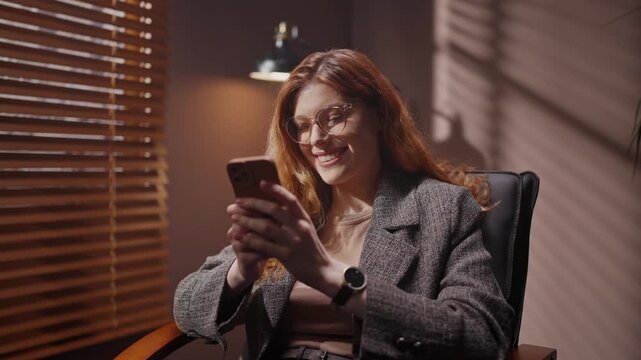 Red head woman looking at smartphone screen. Female isolated in dark room. Woman wearing jacket and smiling. Female sitting on chair isolated in dim room. Woman in glasses satisfied with chatting.