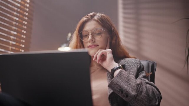 Focused view of woman working with laptop. Red head woman using laptop late in evening. Female sitting on chair isolated in dim room in loft style. Woman wearing glasses and smiling at screen.