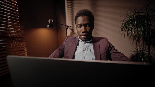 Front view of African American man working with laptop. Male using laptop late in evening. Professional man wearing suit looking at laptop screen. Office manager working in dim room. Ethnic diversity.