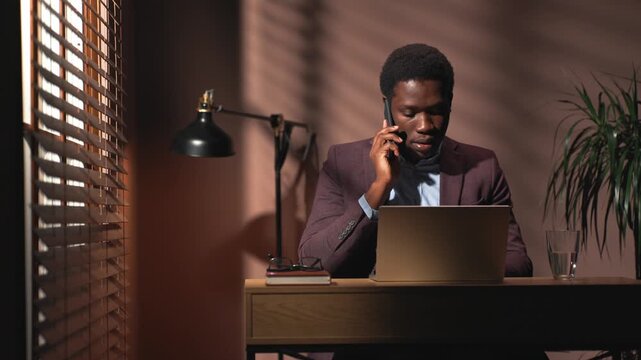 African American man sitting at office table with lamp. Man in suit sitting near window with blinds. Male conducting business call in dark office room. Company director sitting in front of laptop.