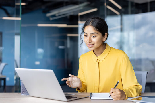 Young Indian woman talking on video call on laptop, sitting in office chair and taking notes in notebook