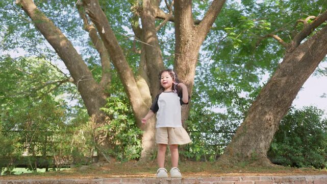 A young toddler girl dances playfully on a brick ledge in a park, experiencing joyful childhood development and connecting with nature during outdoor recreation.