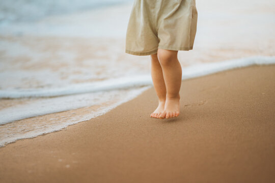 Little Asian toddler girl walking barefoot on a sandy beach watching sea waves during a summer family vacation. Close-up of child's feet on the shore. Concept of childhood discovery.
