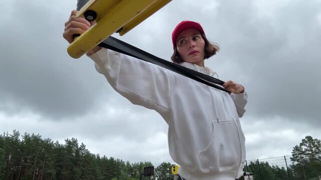 Low angle view of a focused brunette woman in a bright red cap performing an outdoor resistance band chest press workout pulling the elastic loop from a high anchor point