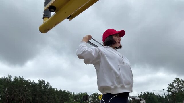 Low angle view of a focused brunette woman  in a bright red cap performing an outdoor overhead triceps extension workout with a black elastic resistance band anchored high