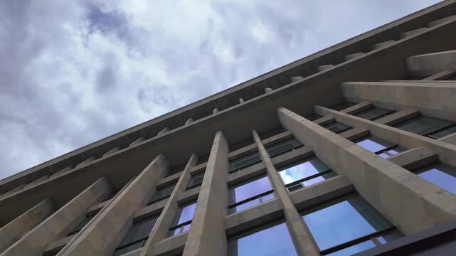 Dramatic low-angle view of a modern brutalist building with a concrete geometric facade and reflective blue windows, showing imposing and rhythmic architectural patterns against a cloudy sky