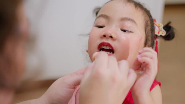 A young Asian girl in a traditional red dress gets festive makeup and lip gloss applied by her mother, preparing for a Chinese New Year family celebration.
