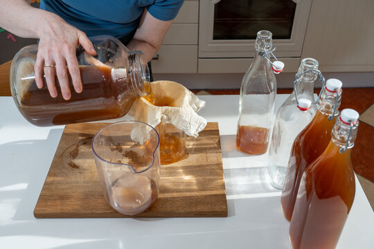 High-angle perspective of a woman decanting a large glass vessel of homemade kombucha through a cotton filter, separating the liquid from the SCOBY for bottling.