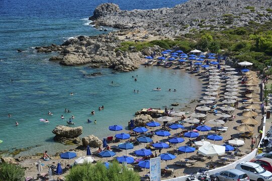 Beach full of deckchairs and blue umbrellas, lots of people in and around the clear water, surrounded by rocks, Anthony Quinn Bay, Vagies Bay, Rhodes, Dodecanese, Greek Islands, Greece