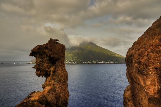 Stromboli island seen from the offshore islet of Strombolicchio, dramatic rock formation with volcanic island in the background under a cloudy sky, UNESCO World Heritage Site, Aeolian Islands, Aeolian Islands, Sicily, Italy