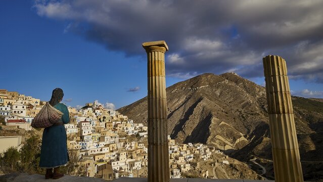 A life-size doll of a woman looks at the landscape framed by ancient columns, with a village in the mountains, Colourful mountain village, Morning light, Olymbos, Karpathos, Dodecanese, Greek Islands, Greece