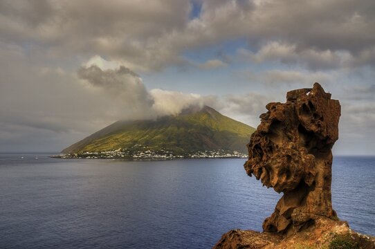 Stromboli island seen from the offshore islet of Strombolicchio, impressive rock in the foreground with volcanic island and clouds in the background, UNESCO World Heritage Site, Aeolian Islands, Aeolian Islands, Sicily, Italy