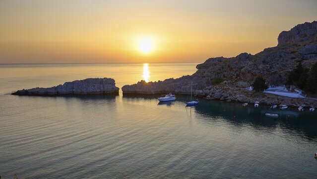 Golden glowing sunset over the calm sea next to rocky cliffs, Paulus Bay, below the Acropolis of Lindos, Lindos, Rhodes, Dodecanese, Greek Islands, Greece