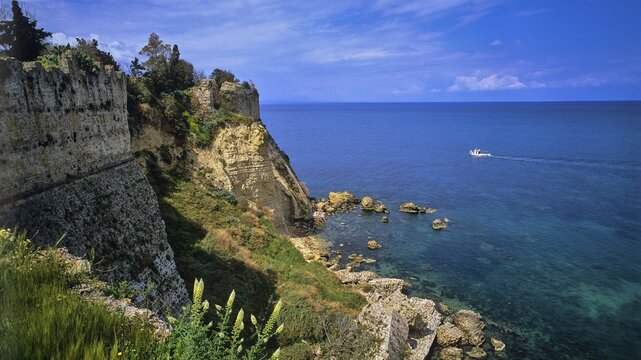 Steep cliffs and old fortress walls overlook the clear blue sea under a partly cloudy sky, sea fortress Methoni, Peloponnese, Greece
