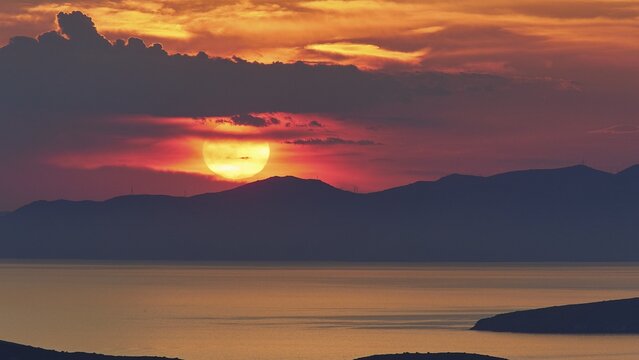 Sunset, sun large under grey clouds and directly above a mountain range, headlands as silhouette in foreground, sea calm, sky orange-red, Palaiopolis, Andros Island, Cyclades, Greece