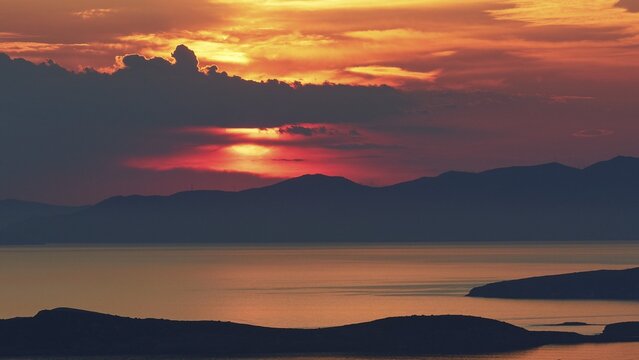 Sunset, sun big behind grey clouds and over a mountain range, headlands as silhouette in foreground, sea calm, sky orange-red, Palaiopolis, Andros Island, Cyclades, Greece