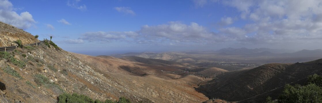 Panorama shot, road, curve, palm trees, Mirador Morro Velosa, barren hilly landscape, blue sky, white clouds, Fuerteventura, Canary Islands, Spain
