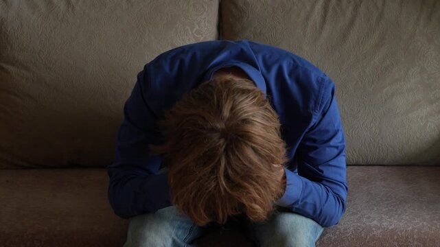Young man (zoomer) with messy hair leaning forward
 and holding his head. Visibly troubled, feeling stress, burnout and mental exhaustion. Depression and mental health concept.