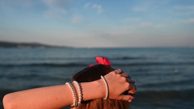 Young woman with long hair wearing pearl bracelets and a shell necklace by the sea during golden hour. A calm summer lifestyle scene with warm sunlight, elegant accessories, and a relaxed 