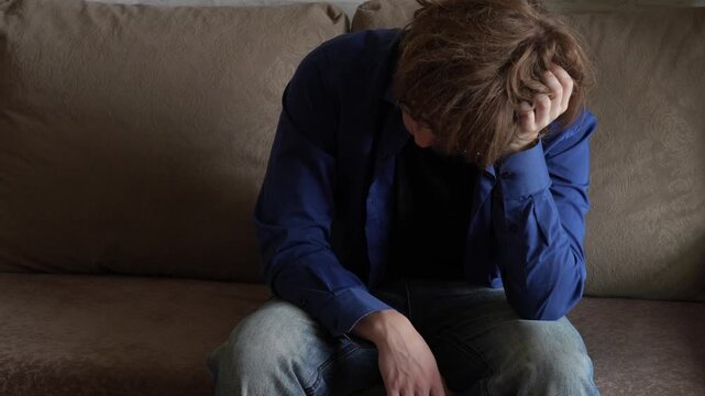 Young man (zoomer) with messy hair sitting
 and holding his head. Visibly troubled, feeling stress, burnout and mental exhaustion. Depression and mental health concept.