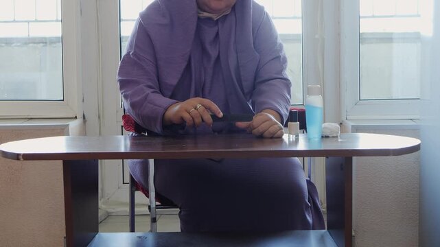 A woman in a lilac dress is doing her manicure and filing her nails