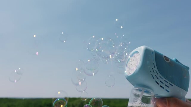 A hand holds a blue plastic bubble machine against a clear sky. Soapy foam gathers at the nozzle of the toy device. A distant green forest completes the sunny outdoor summer landscape.