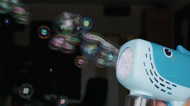 A blue whale-shaped toy bubble blower shoots a continuous stream of iridescent glowing bubbles in a dimly lit room. Concept of childhood joy, summer fun, outdoor play, and imagination.