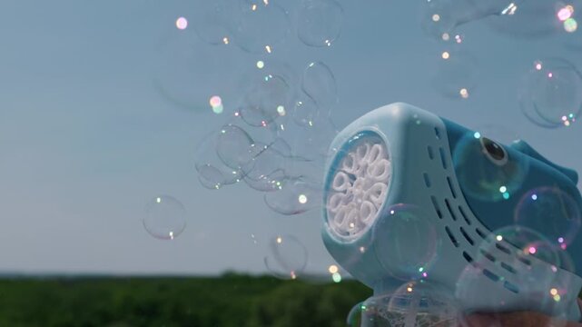 A blue shark toy bubble machine blows a stream of iridescent soap bubbles into the air. The bright clear blue sky and green landscape create a cheerful summer atmosphere outdoors.