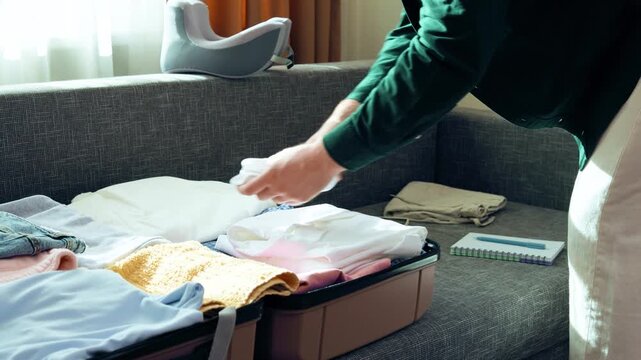 Man doing travel preparation for a summer vacation. He is packing a suitcase with clothes on a sofa and checking a packing list to complete his baggage preparation. High quality 4k footage