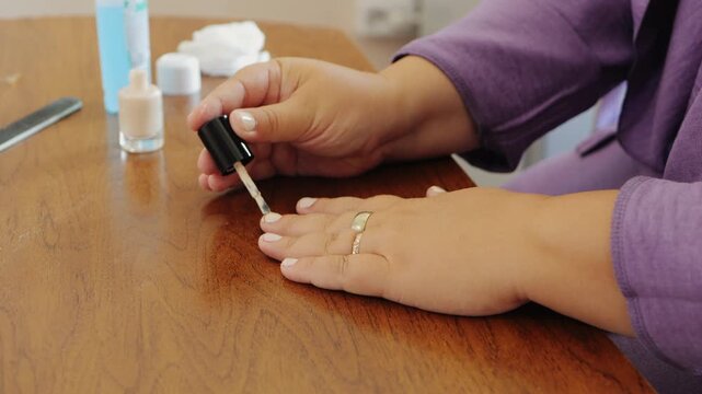 A woman paints her nails with light polish at a table