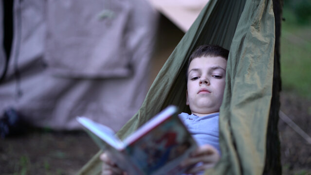 A young boy holds an open book while resting in a green hammock outdoors during summer. Calm lifestyle footage of relaxed child in peaceful natural setting.
