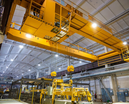 A massive yellow overhead bridge crane with an operator's cabin and heavy-duty lifting hooks spans across the ceiling of a large industrial manufacturing factory workshop.
