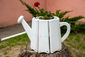 A decorative white wooden container shaped like a watering can stands on a tree stump, holding a blooming red geranium flower in an outdoor garden setting. © Dmitry