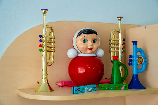 A traditional red Soviet nevalyashka roly-poly doll stands on a wooden shelf surrounded by colorful plastic toy trumpets and harmonicas against a blue wall.
