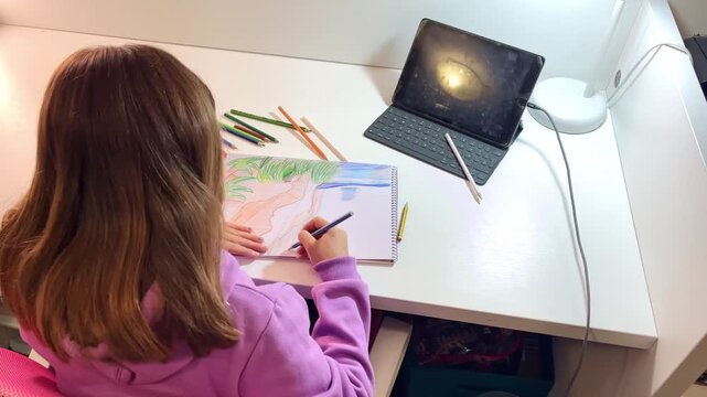 High angle shot of a young girl sitting at a white desk in her bedroom, using color pencils to sketch a beach landscape in her notepad while using a digital tablet screen as a creative reference