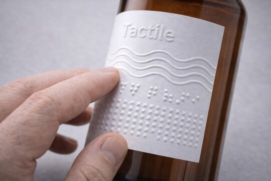 Tactile Braille Label on Glass Bottle. Closeup of hand touching embossed tactile label with braille text on brown glass bottle for accessibility.